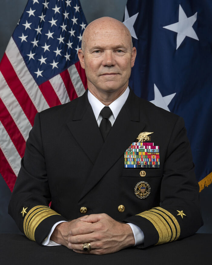 Pete Hegseth in military uniform with medals, seated in front of American and blue flags, stern expression.