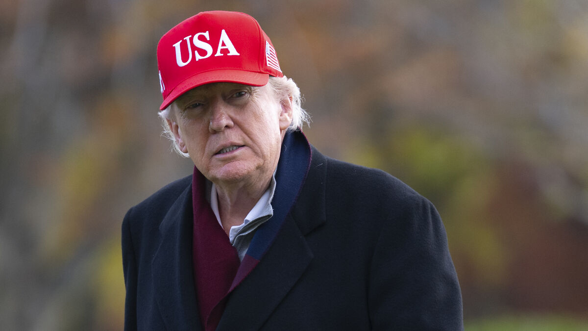 Former President Trump wearing a red USA cap and coat outdoors amid blurred autumn colors related to White House MRI scan news.