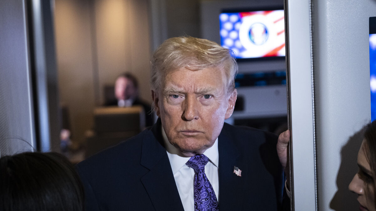Donald Trump in a suit and purple tie, looking serious while interacting with reporters in a crowded room.