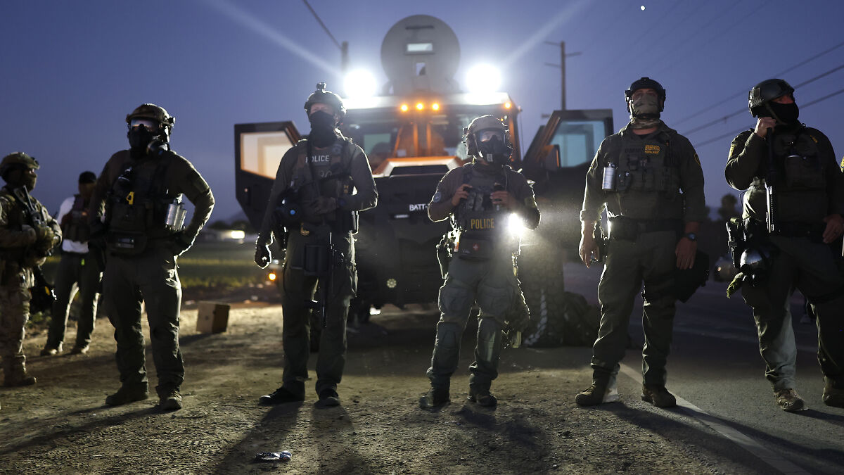 ICE officers in tactical gear standing in front of an armored vehicle during a nighttime raid operation.