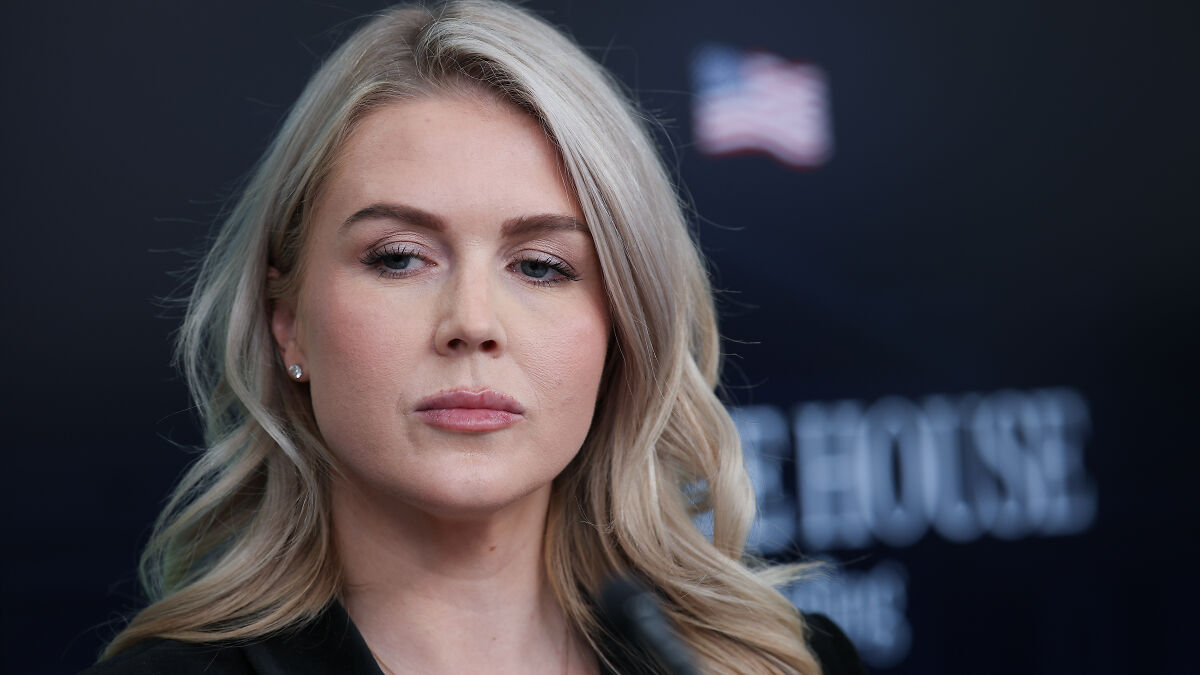 Karoline Leavitt with a serious expression at a podium, with an American flag in the blurred background.