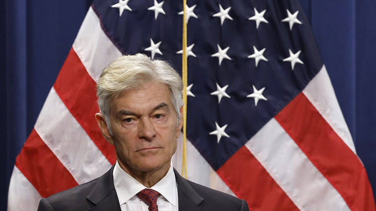Dr. Oz speaking seriously in front of American flags, addressing federal workers after government shutdown advice.