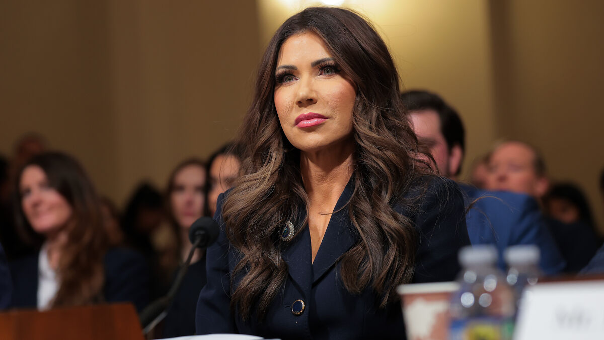 Kristi Noem testifying at a hearing, focused and attentive with blurred audience members in the background.