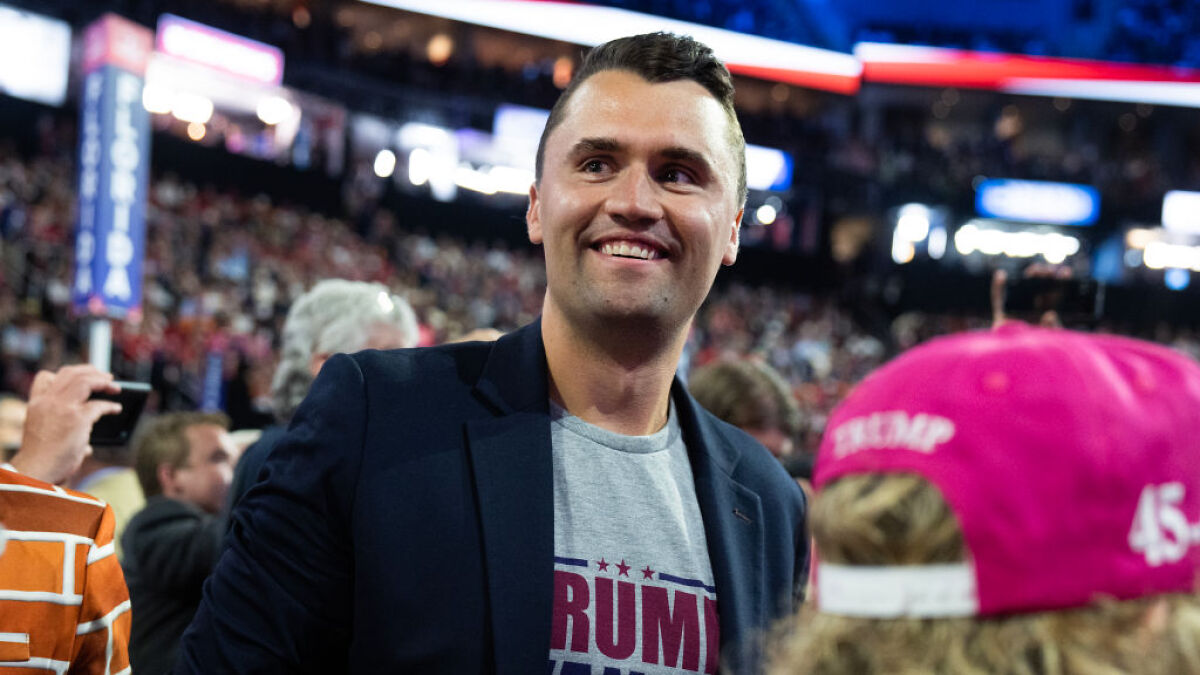 Charlie Kirk at a crowded event, smiling and wearing a blazer over a Trump campaign shirt.