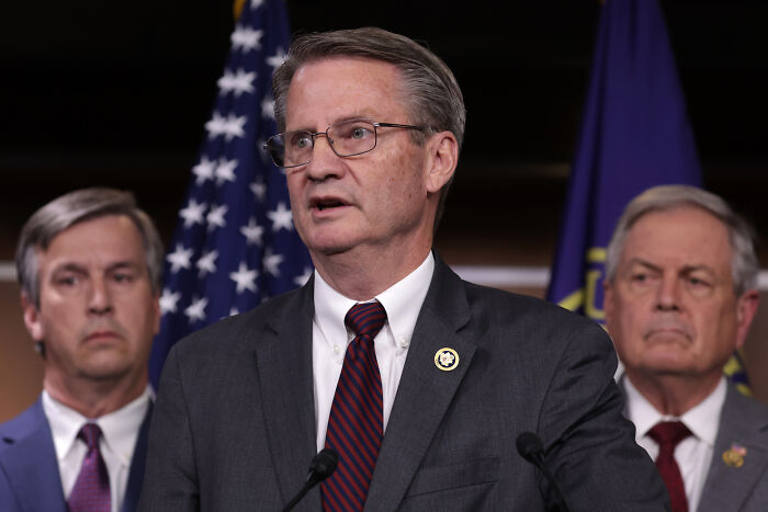 A man in a suit and glasses speaking at a podium with American and dark blue flags behind him, discussing Taliban funding. A man in a suit and glasses speaking at a podium with American and dark blue flags behind him, discussing Taliban funding.