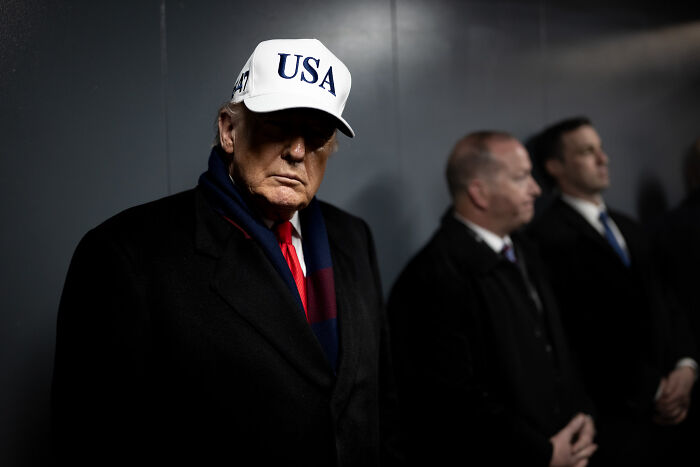 Former president wearing USA hat and dark coat, with two men blurred in background during a staged TV address setting. Former president wearing USA hat and dark coat, with two men blurred in background during a staged TV address setting.