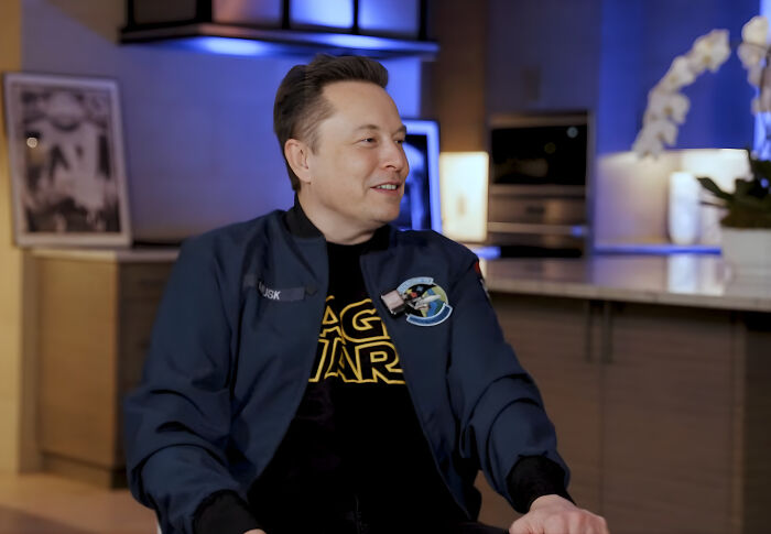 Elon Musk seated in a casual setting wearing a Space Wars shirt and dark jacket, engaged in conversation. Elon Musk seated in a casual setting wearing a Space Wars shirt and dark jacket, engaged in conversation.