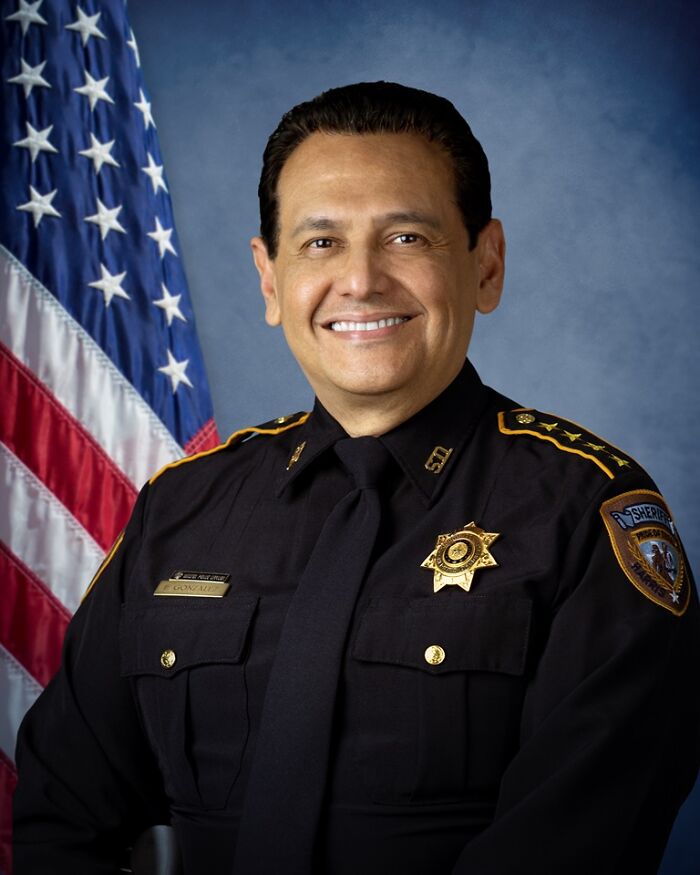 Texas sheriff smiling in uniform with badge and American flag in background representing law enforcement response. Texas sheriff smiling in uniform with badge and American flag in background representing law enforcement response.