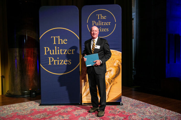 Man at Pulitzer Prizes backdrop holding folder, image referencing Trump and NYT Pulitzer photographer Man at Pulitzer Prizes backdrop holding folder, image referencing Trump and NYT Pulitzer photographer