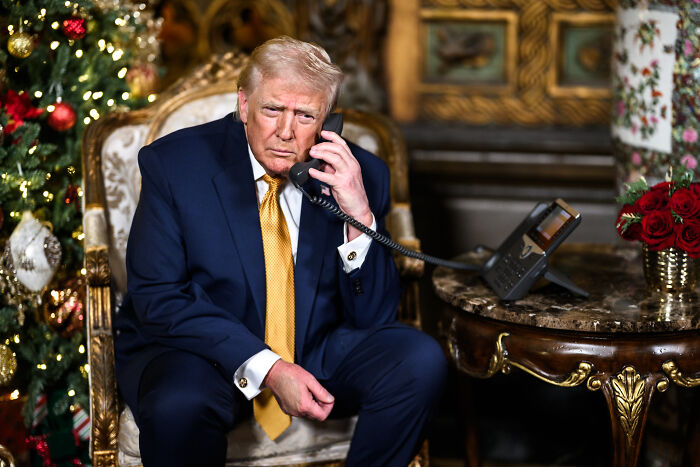 Former President Donald Trump in a suit sitting while holding a phone during a decorated holiday setting with Christmas tree and red roses.