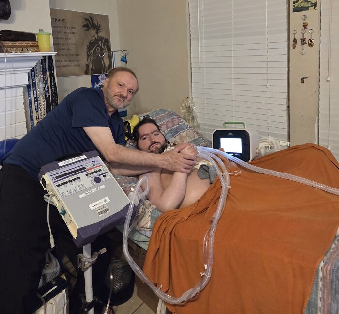 Father comforting disabled son in bed with medical equipment nearby, highlighting ICE blocking funeral attendance.