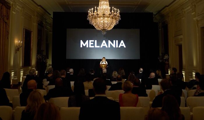 Melania Trump standing on stage at a documentary screening event with an elegant chandelier overhead and seated audience. Melania Trump standing on stage at a documentary screening event with an elegant chandelier overhead and seated audience.