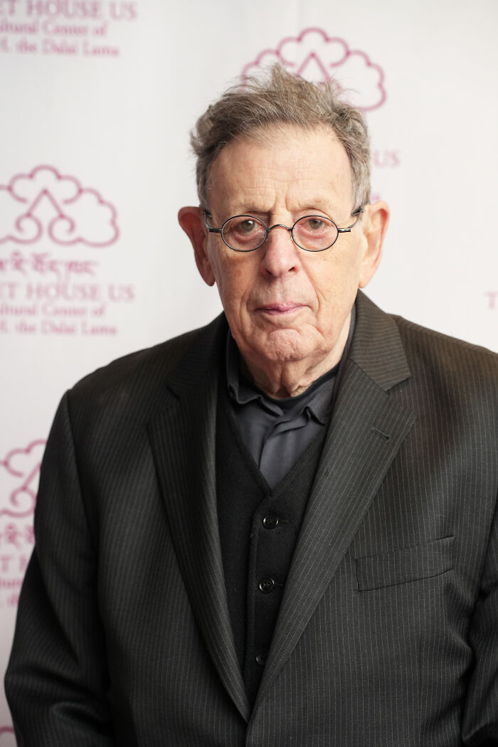 Philip Glass wearing glasses and a pinstripe suit, posing at an event related to iconic composer and symphony news.