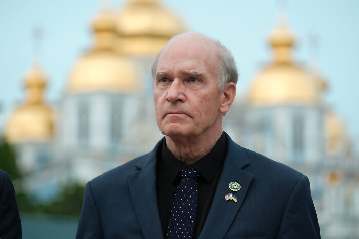 Male lawmaker in suit with NATO territory and Greenland threats backdrop during a formal outdoor event.