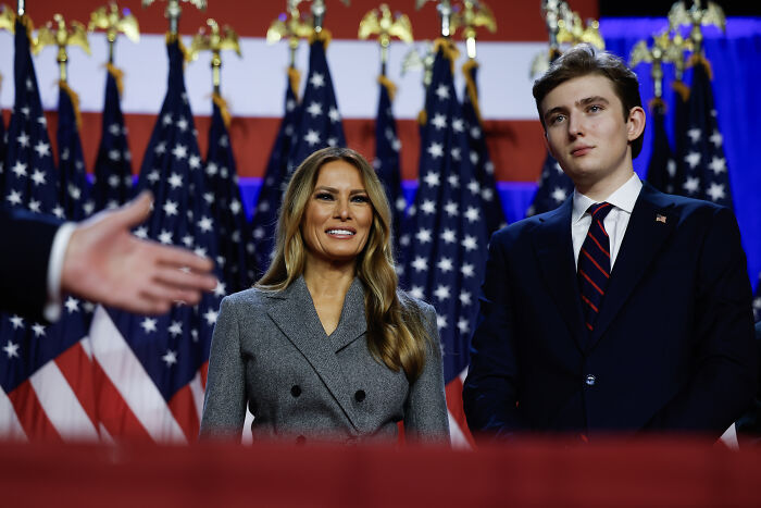 Melania Trump and Barron Trump standing in front of multiple American flags at a formal event. Melania Trump and Barron Trump standing in front of multiple American flags at a formal event.