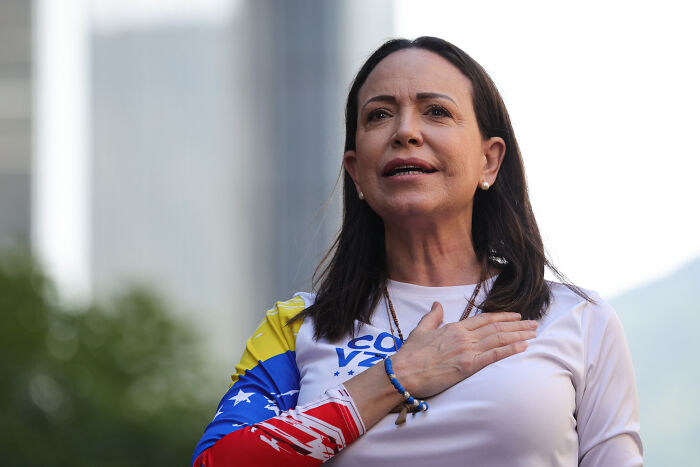 Woman with hand on chest wearing a colorful long sleeve shirt, representing Trump welcoming Machado&rsquo;s peace prize offer.