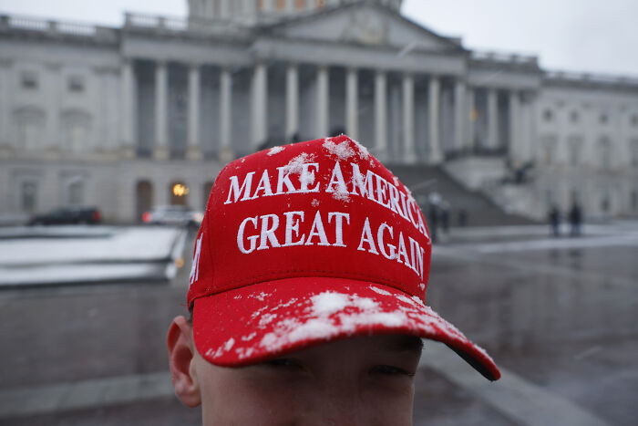 Red Make America Great Again hat worn by a person with snow on it, in front of a large government building.