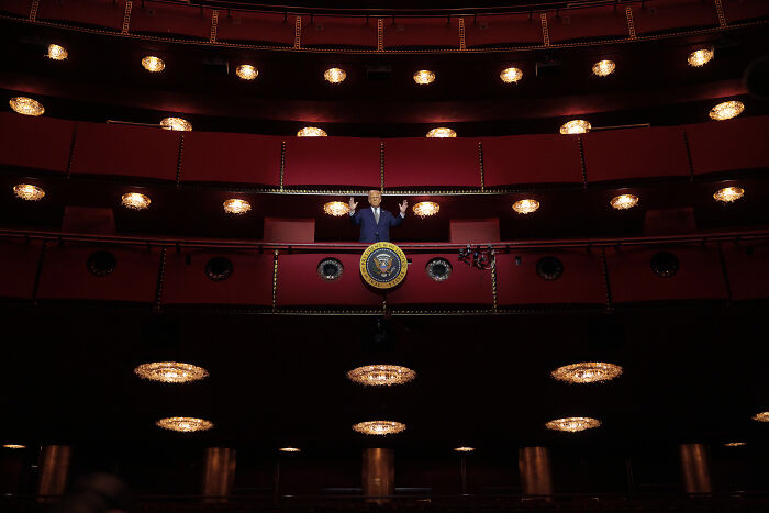 Man standing in a red theater balcony with presidential seal, symbolizing iconic composer Philip Glass pulling symphony from Kennedy Center.