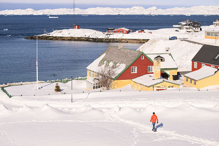 Colorful houses covered in snow near the shore of Greenland with a person walking and a clear blue sky above.