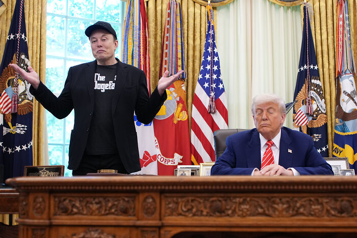 Elon Musk standing with arms outstretched near former President Trump seated at Resolute Desk in Oval Office with flags behind.