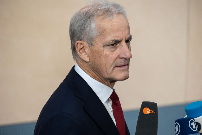 Older man in suit speaking to reporters holding microphones about Trump and peace issues in a formal setting