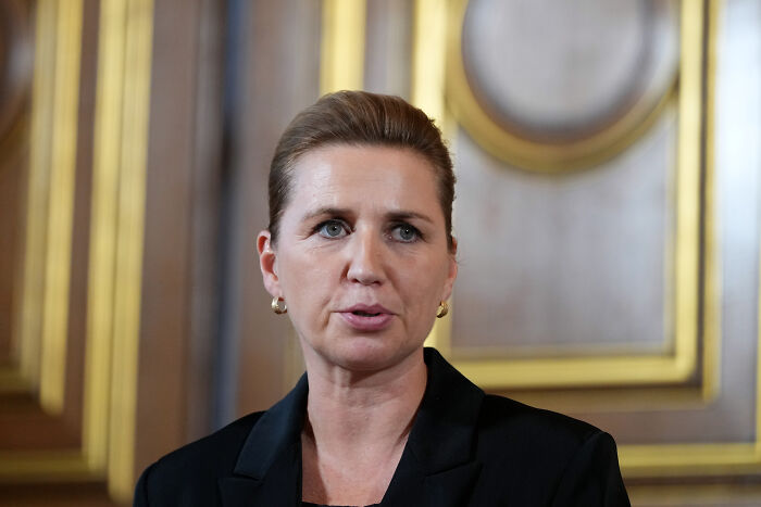 A woman in a black blazer speaking in a formal room with ornate gold and wood-paneled walls.