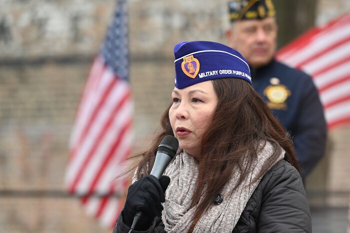 Woman in military hat speaking into microphone at outdoor event with American flags, related to combat veteran controversy. Woman in military hat speaking into microphone at outdoor event with American flags, related to combat veteran controversy.