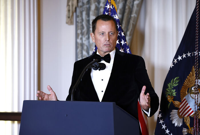 Kennedy Center President speaking at a podium in a tuxedo with American flags behind during a formal event.
