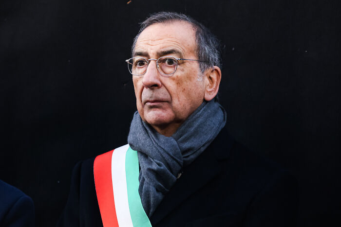 Man wearing glasses, dark coat, and scarf with an Italian flag sash, looking serious against a black background. Man wearing glasses, dark coat, and scarf with an Italian flag sash, looking serious against a black background.