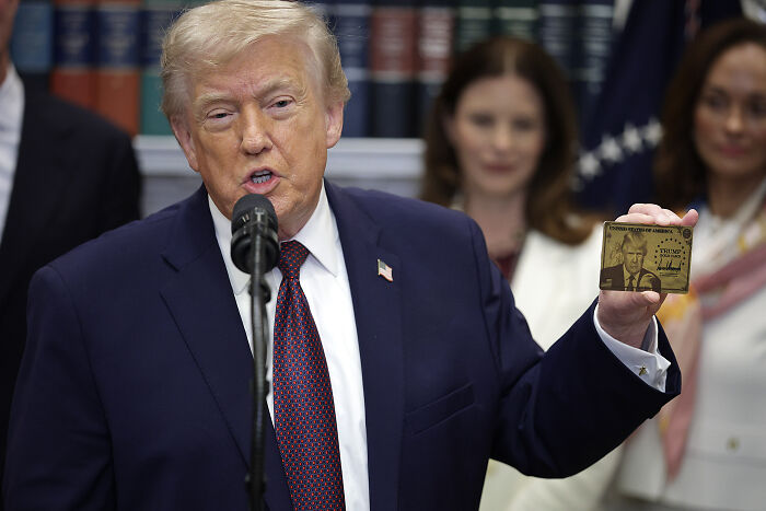 Donald Trump wearing a suit and tie, speaking at a microphone, holding a gold card with his image and signature. Donald Trump wearing a suit and tie, speaking at a microphone, holding a gold card with his image and signature.