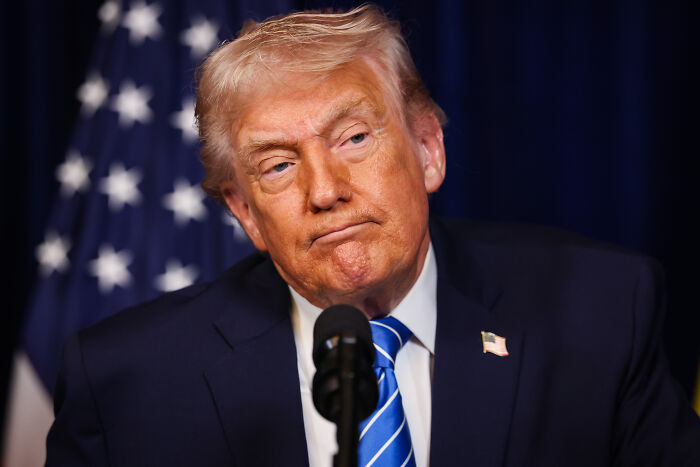 Donald Trump speaking at a podium with an American flag behind him, mentioning Greenland's psychological importance.