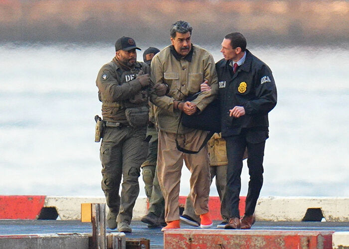 I don't know who this is. Handcuffed man escorted by federal agents on a dock, wearing tan jacket and orange shoes