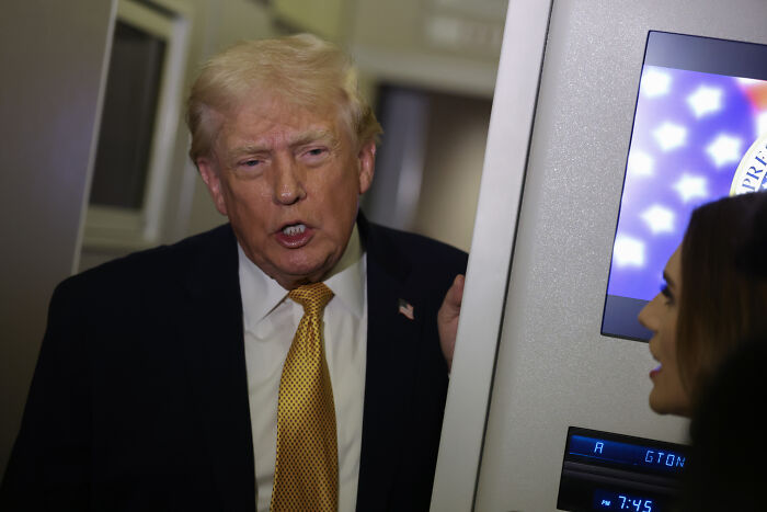 Donald Trump speaking seriously to a woman, with presidential seal visible on a screen in the background.