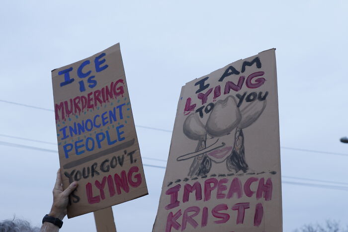 Protest signs addressing ICE accusations and calls to impeach Kristi Noem over controversial statements.