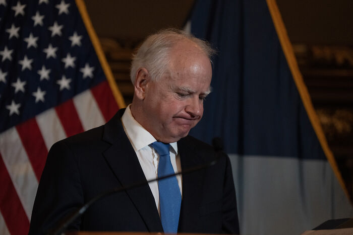 JD Vance at podium, wearing suit and blue tie, appearing somber with American flag and state flag behind him.