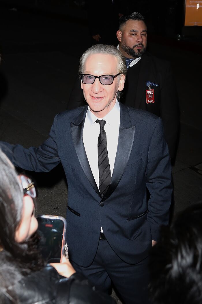 Bill Maher in a dark suit and glasses, reacting unexpectedly as actors honor Renee Good at the Golden Globes event.