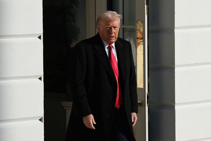 Former President Trump in black coat and red tie leaving a building amid lawmakers blocking invasion of NATO territory.