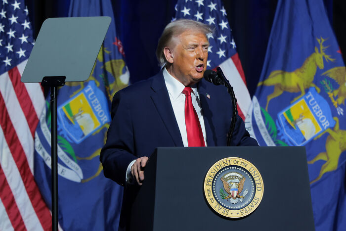 Donald Trump speaking at a podium with presidential seal, captured mid-gesture during a public speech.