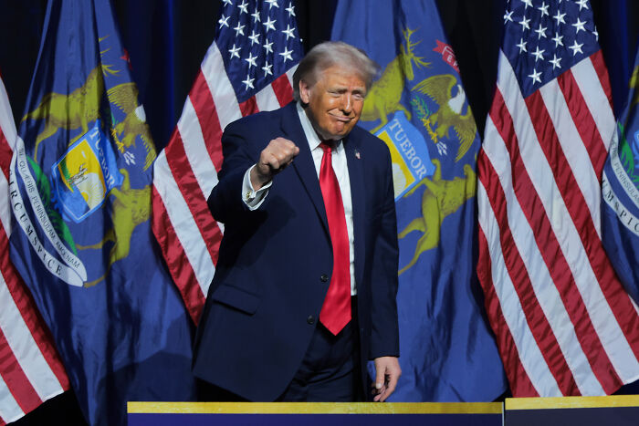 Donald Trump on stage, wearing a suit with red tie, gesturing during a speech with American flags in the background. Donald Trump on stage, wearing a suit with red tie, gesturing during a speech with American flags in the background.