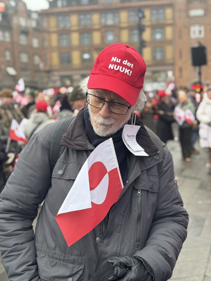 Greenland resident wearing a red hat with Nuuk text and holding the Greenland flag during an outdoor event.