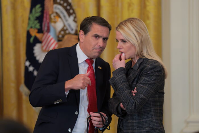 Two people in formal attire engaged in a serious conversation with presidential seal and American flag in background