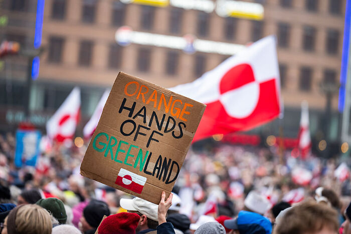 Protesters holding a cardboard sign in a crowd with Greenland flags representing a viral Greenland Reddit post message. Protesters holding a cardboard sign in a crowd with Greenland flags representing a viral Greenland Reddit post message.