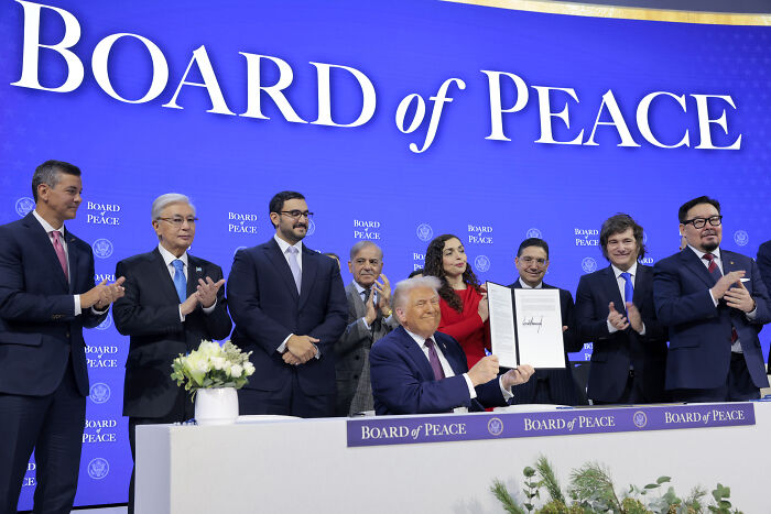 Donald Trump seated at a signing ceremony, surrounded by applauding officials under a Board of Peace banner.