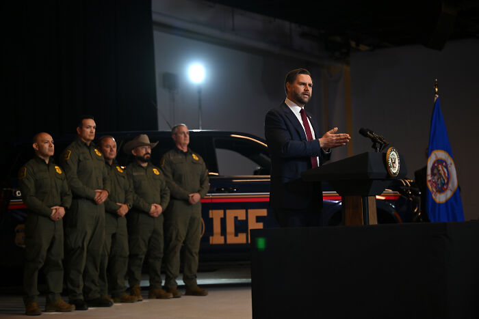 JD Vance speaking at a podium with ICE agents standing beside an ICE vehicle during a press event.