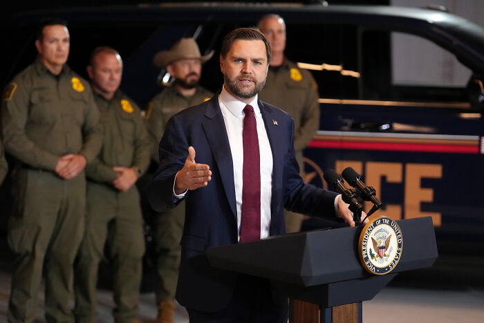 JD Vance speaking at podium with law enforcement officers standing behind during a controversial hearing event. JD Vance speaking at podium with law enforcement officers standing behind during a controversial hearing event.