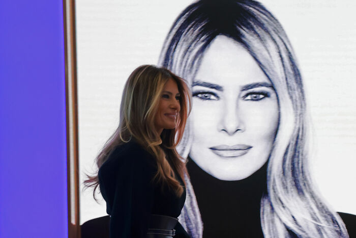 Melania Trump standing near a large black and white portrait of herself at a public event. Melania Trump standing near a large black and white portrait of herself at a public event.