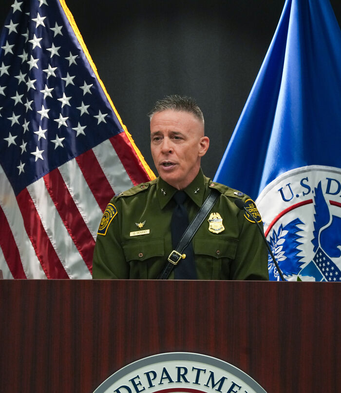 U.S. Border Patrol officer in uniform speaking at a podium with American and Department of Homeland Security flags behind him