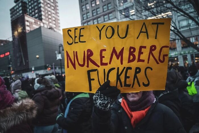 Protester holding an anti-ICE sign at a crowded demonstration in a city setting during dusk.