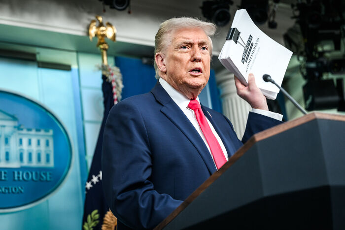 Donald Trump at a podium holding documents, speaking on a covert plan to deploy ICE agents during the Olympics. Donald Trump at a podium holding documents, speaking on a covert plan to deploy ICE agents during the Olympics.