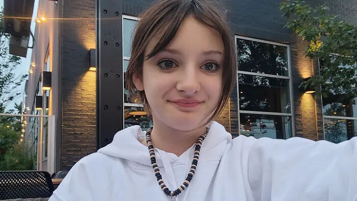 Young girl wearing a white hoodie and beaded necklace, outdoors at dusk, honoring children lost in Canada school attack.